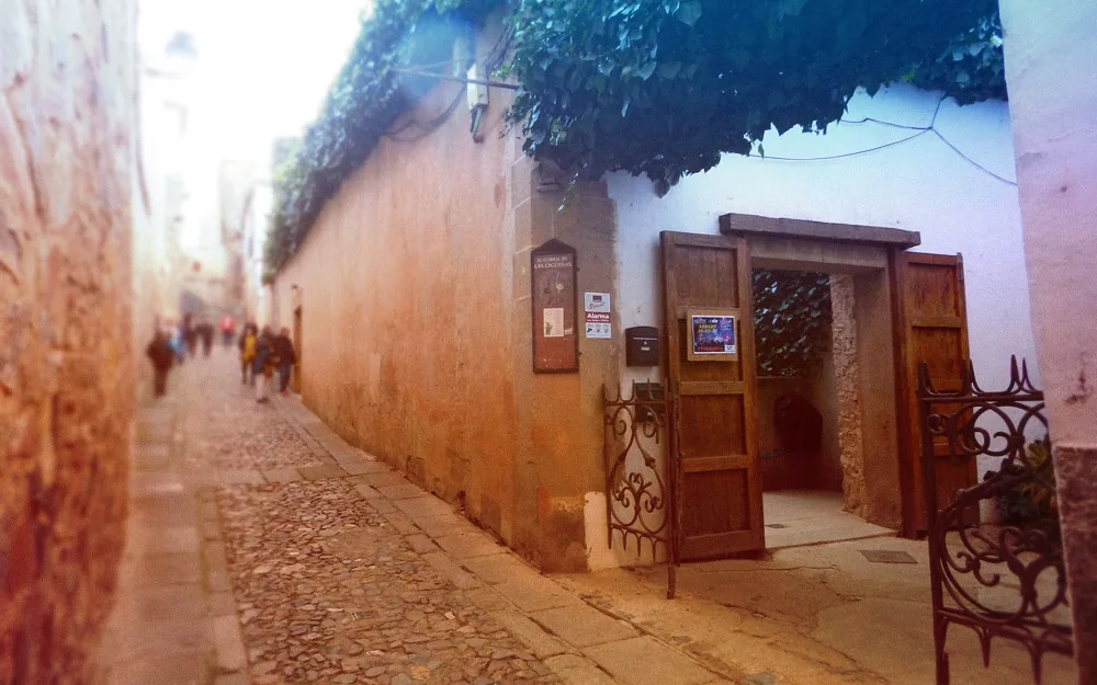 Entrada principal de El Corral de las Cigüeñas, en la Cuesta de Aldana: puerta de madera abierta en el muro de piedra, con hiedra sobre la fachada