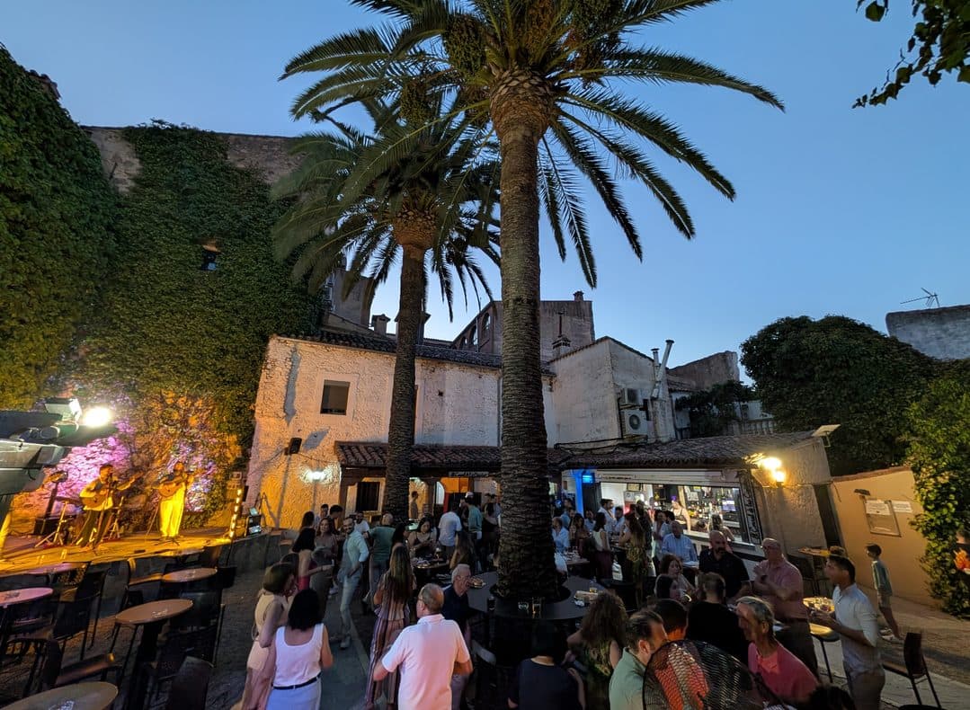Patio de El Corral de las Cigüeñas lleno de gente al anochecer, con las palmeras, el muro de hiedra y un dúo tocando en el escenario
