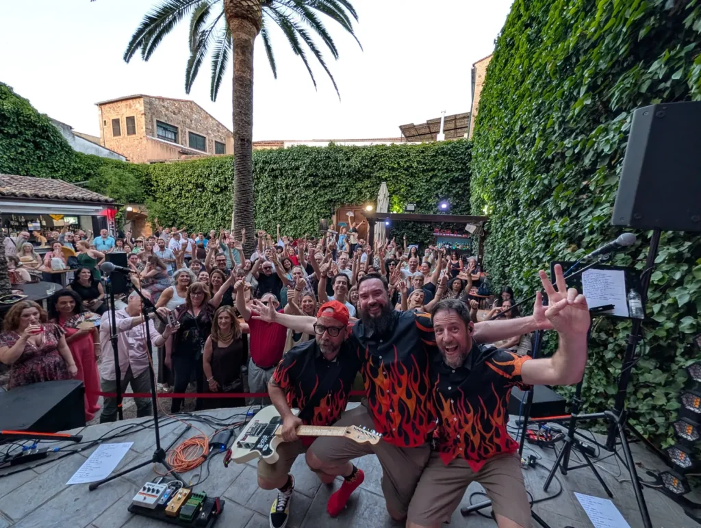Tres músicos con camisas de llamas posan desde el escenario con el patio lleno de gente aplaudiendo detrás