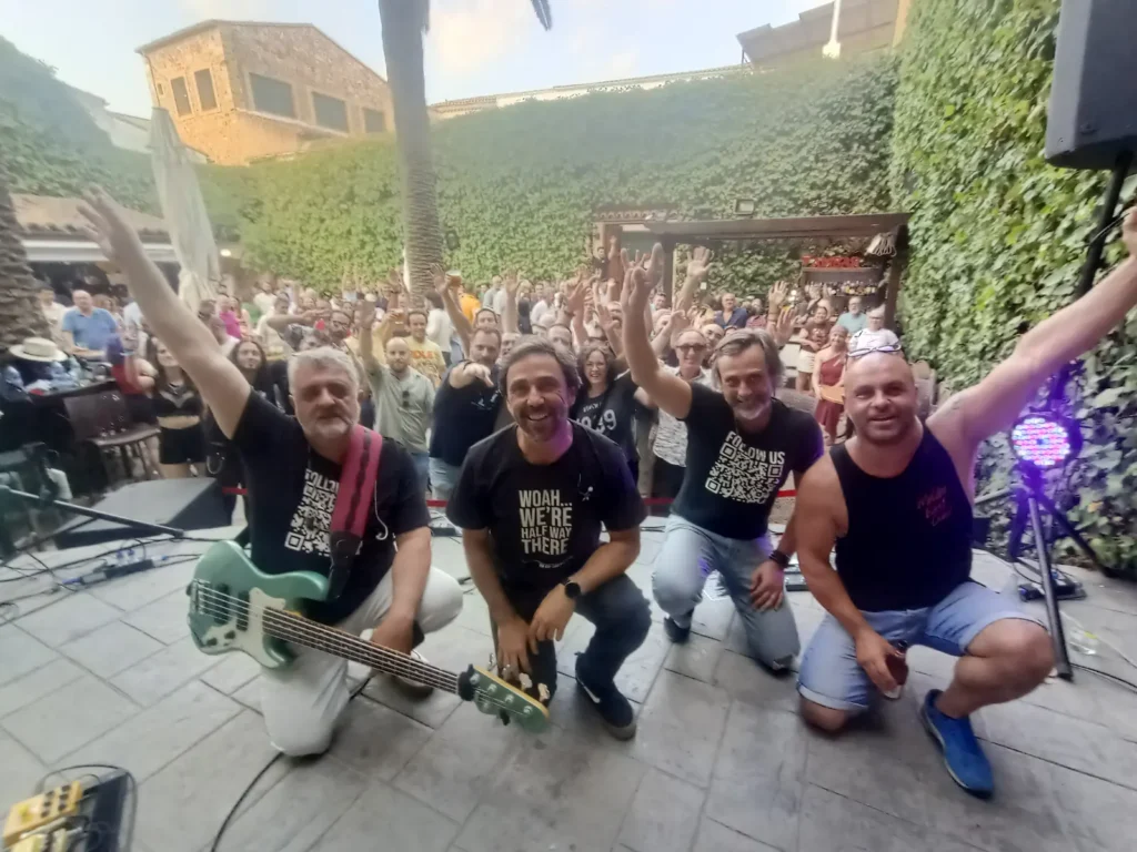 Los cuatro músicos de una banda posan de rodillas con el brazo en alto y el patio lleno de público detrás