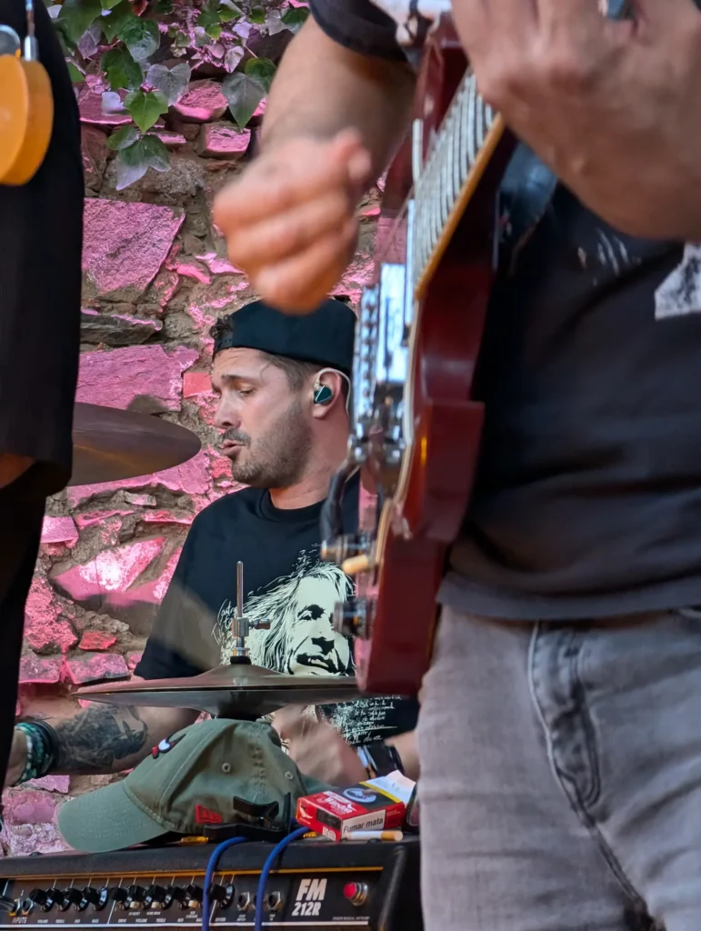 El batería tocando, visto entre los brazos del guitarrista, con el muro de piedra iluminado de rosa
