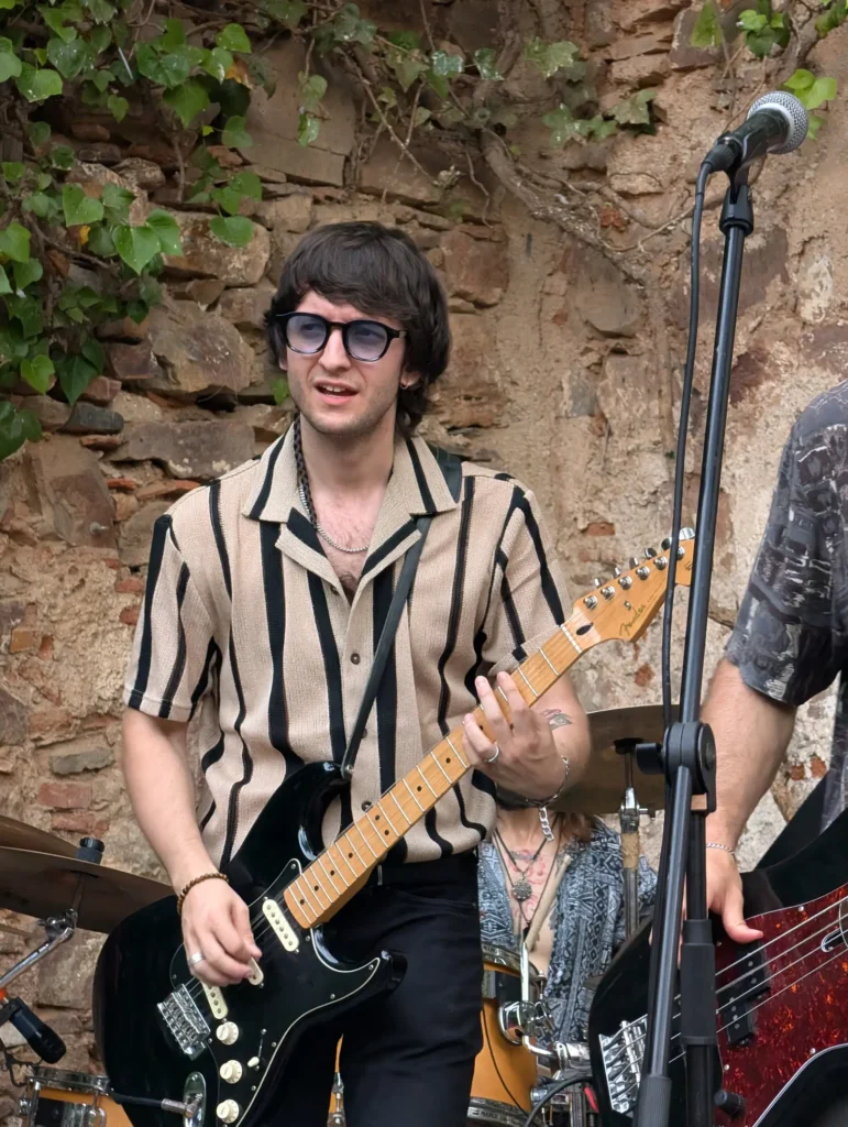 Un guitarrista joven con gafas de sol y camisa de rayas toca su Stratocaster ante el muro de piedra