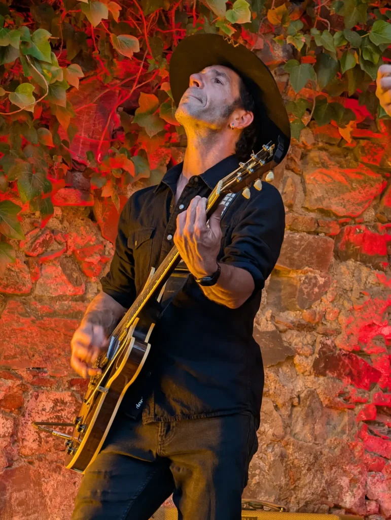 Un guitarrista con sombrero toca mirando hacia arriba, ante el muro de hiedra y piedra iluminado de rojo