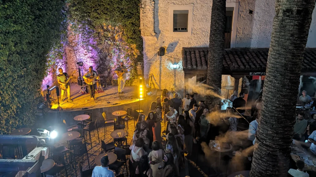 El patio de noche, con el escenario iluminado y el público entre las palmeras
