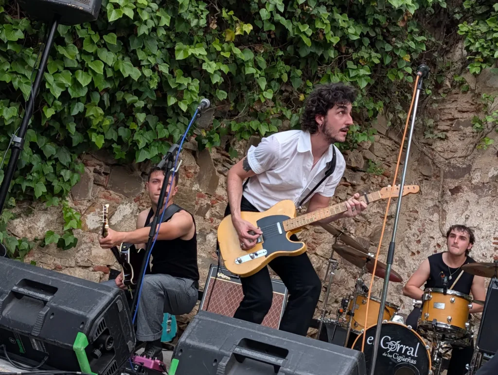 Un trío de rock tocando en el escenario del patio ante el muro de hiedra, con el bombo de El Corral